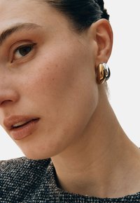 Guld- och silverörhängen med slät, metallisk yta, mot en strukturerad svartvit klädsel.