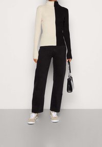 Rippstrick-Turtleneck-Oberteil in Schwarz und Beige, halb-halb Design. Kombiniert mit schwarzen weitem Hosen und beigen Sneakers, hält eine kleine schwarze Tasche.