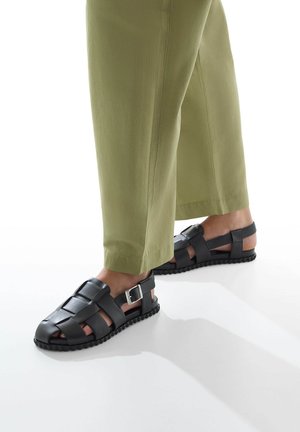 Sandali neri in pelle con design intrecciato, fibbia regolabile e suola in gomma. Abbinati a pantaloni tapered verde oliva.