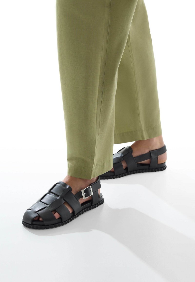 Sandalias de cuero negro con diseño trenzado, hebilla ajustable y suela de goma. Combinadas con pantalones verde oliva, ajustados.