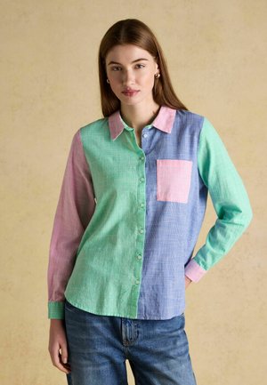 Kvinde iført en skjorte med lange ærmer med pastelfarvede blokke i pink, grøn og blå, kombineret med blå jeans mod en ensfarvet baggrund.