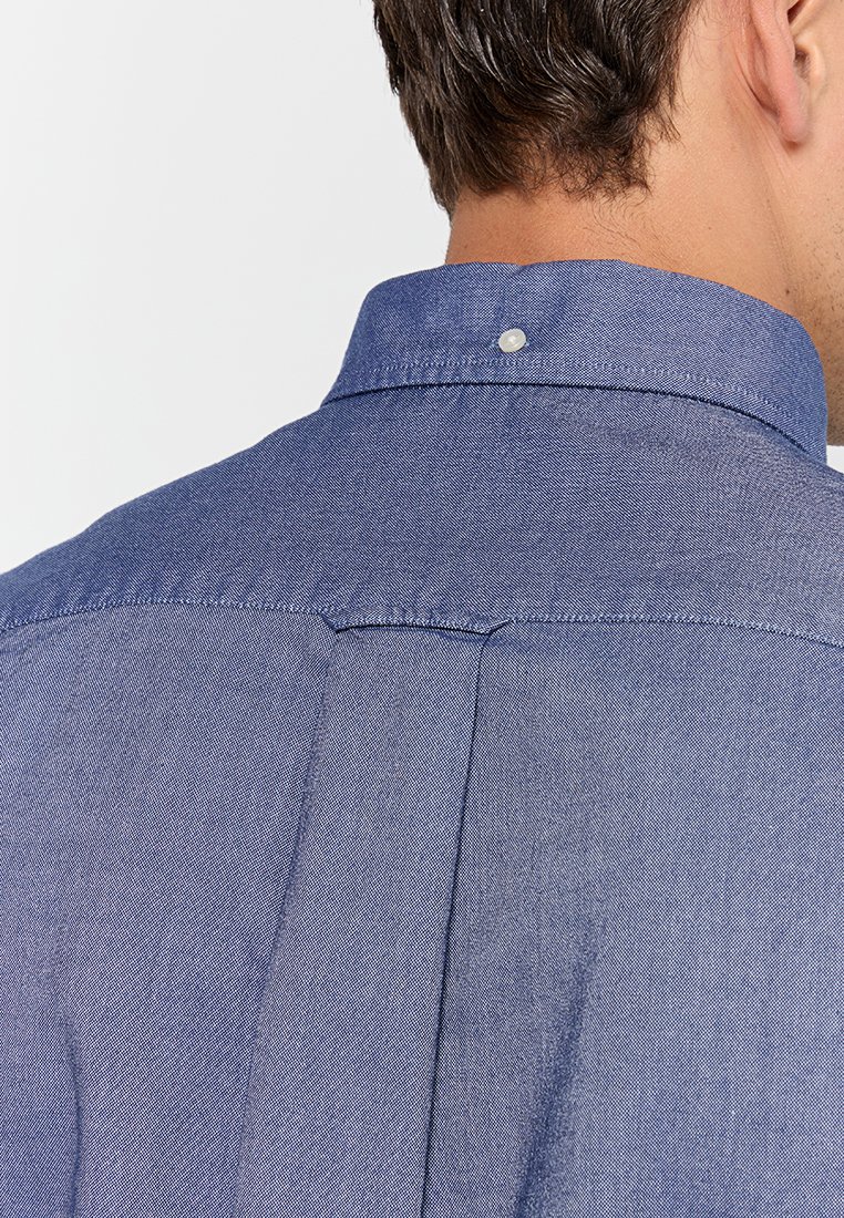 Camisa de mezclilla azul, vista trasera, con cuello abotonado, detalles de costura y una sutil textura con un acabado suave.