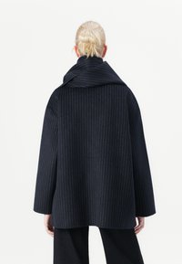Marineblauer Mantel mit vertikalen Streifen, gefertigt aus strukturiertem Stoff. Verfügt über Kragen in Überbreite, oversized Silhouette und herabgesetzte Schultern.