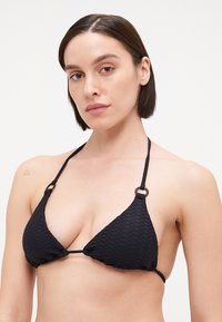 Svart bikini topp med en teksturert, diamantmønster. Har trekantede kopper, slanke stropper og sirkulære hardware-detaljer på skuldrene.