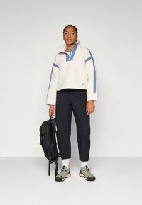 Witte fleece trui met blauwe accenten, halve rits aan de kraag, gecombineerd met een zwarte broek en grijze schoenen. Model draagt een zwarte rugzak.