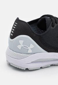 Chaussure de sport noire avec une tige en mesh texturé, des accents gris et un logo blanc. Elle présente une semelle rembourrée et un design respirant pour un bon soutien.