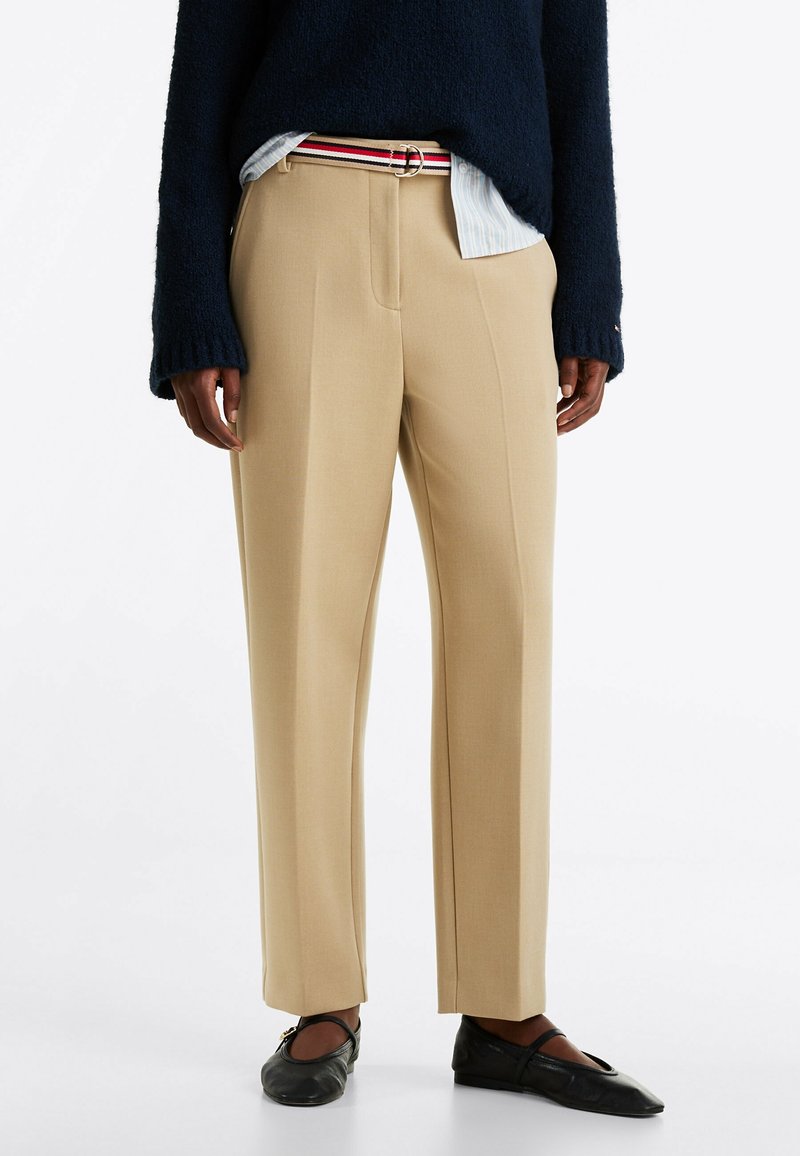 Pantaloni beige su misura con gamba dritta, caratterizzati da pieghe frontali e una texture liscia. Abbinati a un maglione blu navy e ballerine nere.