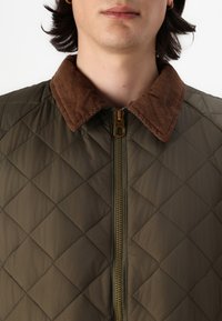 Giacca imbottita verde oliva con colletto in velluto a coste marrone e cerniera dorata. Presenta una texture a motivo diamante e una costruzione leggera.