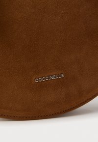 Borsa a mano in camoscio marrone con texture liscia, forma arrotondata e logo "Coccinelle" in argento impresso vicino alla base. Bordi cuciti visibili.