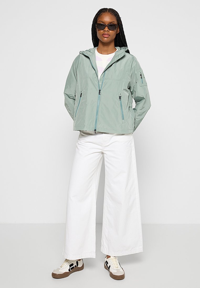 Hellgrüne, leichte Jacke mit Kapuze, versehen mit Reißverschluss-Taschen und atmungsaktivem Material, kombiniert mit weißen weiten Hosen und Sneakers.