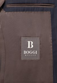 Tumšs jūras zilas jakas iekšpuse, kurai ir brūna auduma rāvējslēdzis ar izšūtu uzplecu, uz kura redzams vārds "BOGGI" un "MILANO" baltā krāsā, akcentēts ar stilizētu "B".