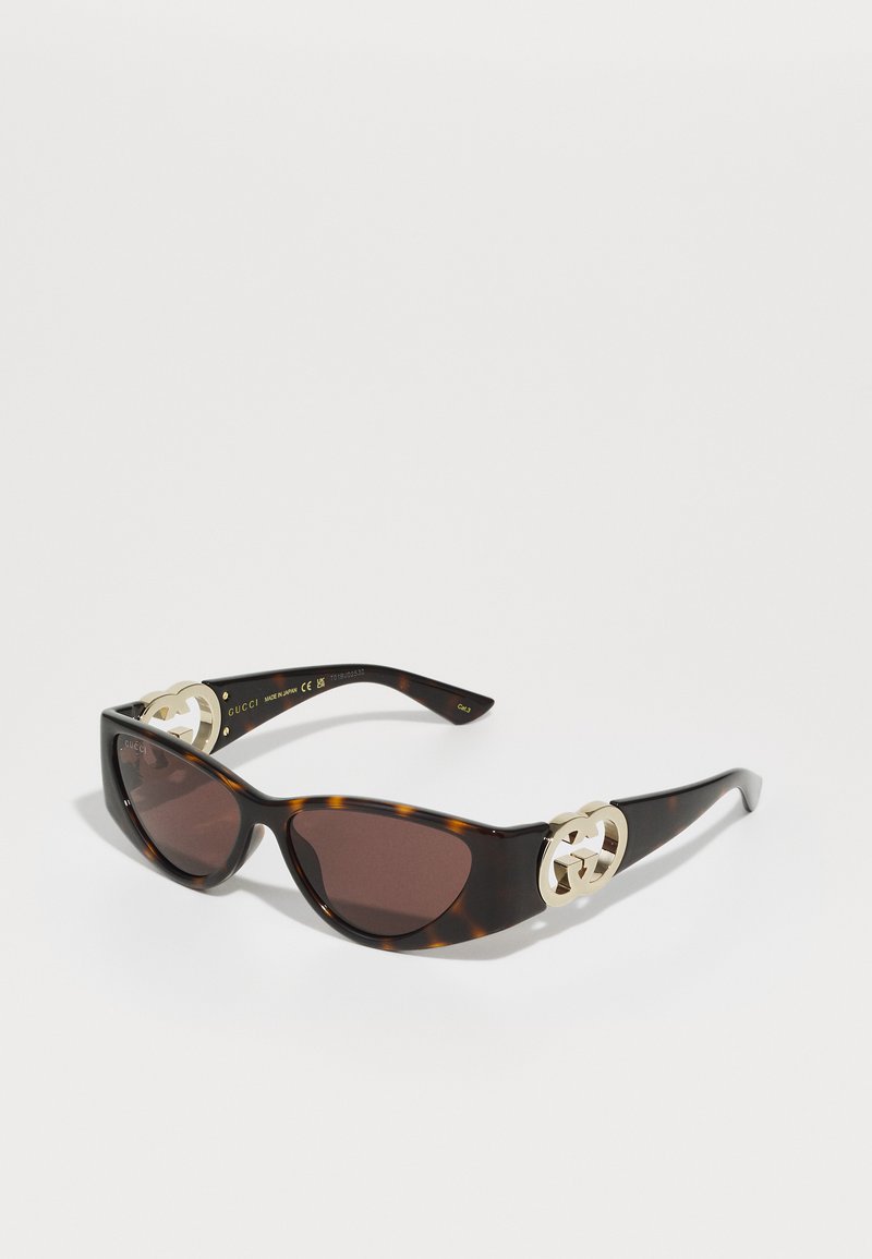 Occhiali da sole Gucci con montatura tartarugata, lenti scure e hardware in oro sulle tempie. Design slanciato e angolare con superficie liscia.