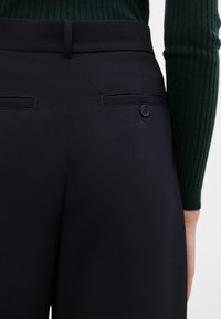 Vue de dos d'une personne portant un pantalon noir sur mesure avec une poche arrière boutonnée et un haut à manches longues côtelé vert foncé.