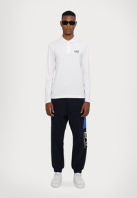 Hvit langermet polo-skjorte med krage og knapper, kombinert med marineblå joggebukser med blå detaljer og grafiske elementer, iført hvite joggesko.
