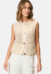 Strikket ærmeløs vest i lys beige med åben vævning, der har to forlommer og en knaplukning med dekorative detaljer.