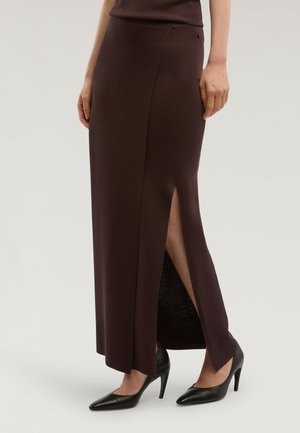 Mujer con una falda larga de color marrón oscuro con una abertura frontal, combinada con zapatos de tacón alto negros de punta afilada, de pie frente a un fondo liso.