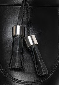 Scarpa di pelle nera con due nappine nere e accenti metallici argentati cilindrici, che mettono in evidenza una superficie liscia.