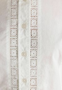 Weiße Bluse mit einer zentralen Knopfleiste, dekorativem Häkelspitzeneinsatz und bestickten Blumenmustern sowie kleinen Knopfverschlüssen.