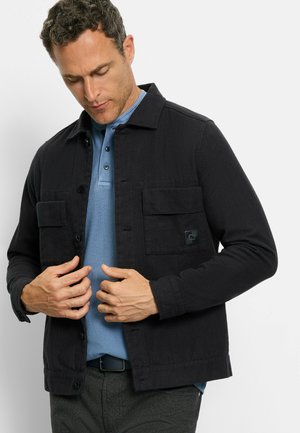 Schwarze Knöpfen-Jacke mit zwei Brusttaschen, strukturiertem Stoff, getragen über einem blauen Polohemd. Enthält ein kleines Logo auf der linken Tasche.