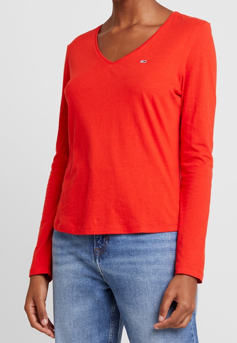 Rode V-hals T-shirt met lange mouwen, gemaakt van katoen met een zachte textuur. Heeft een klein logo op de borst en een ontspannen pasvorm. Draagt blauwe spijkerbroek.