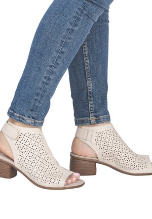 Beine in blauen Jeans, die beige offene Sandaletten mit Absatz, perforierten Blumenmustern und Knöchelriemen tragen.