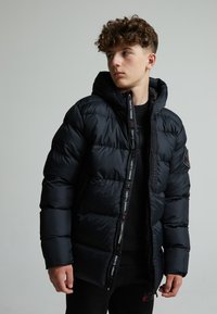 Veste en duvet noire avec capuche, dotée d'une fermeture éclair sur le devant et de poignets élastiques. Elle présente un logo contrasté le long de la fermeture éclair et une texture matelassée.