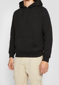 Sudadera negra de tela suave, con bolsillo tipo canguro, capucha con cordón y puños y dobladillo acanalados, combinada con pantalones beige claros.