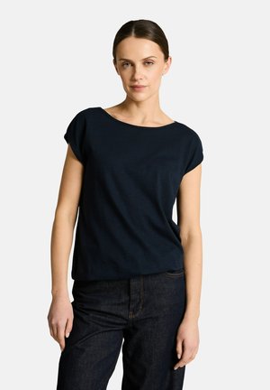 Frau mit dunkelbraunen Haaren, die ein schwarzes kurzärmeliges Oberteil und dunkle Jeans trägt, steht vor einem schlichten hellen Hintergrund.