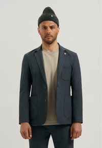 Blazer bleumarin dintr-un material moale, cu două buzunare frontale și un buzunar la piept. Asortat cu o cămașă bej deschis și o căciulă verde închis.