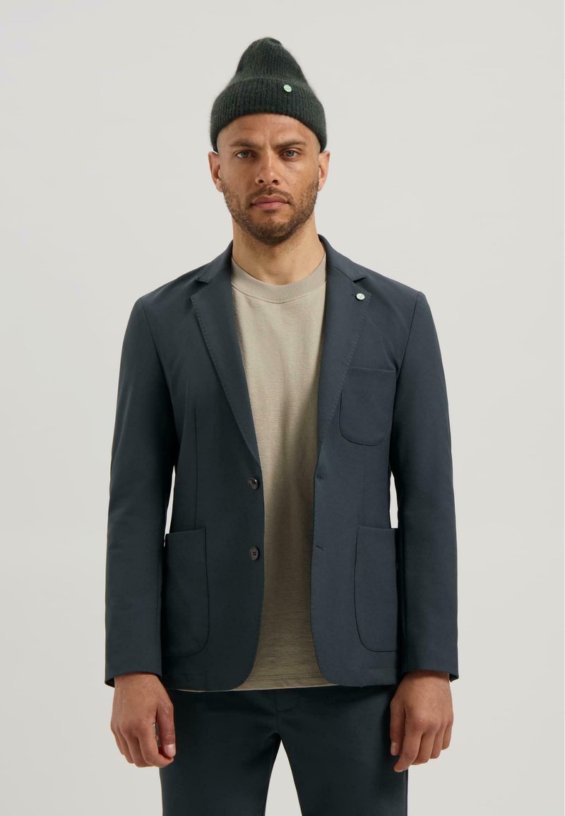 Blazer bleumarin dintr-un material moale, cu două buzunare frontale și un buzunar la piept. Asortat cu o cămașă bej deschis și o căciulă verde închis.
