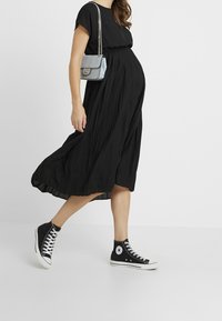 Virallinen musta äitiysmekko lyhyillä hihoilla, laskostettu malli, yhdistetty mustien korkeakorkoisten Converse-tennarien ja pienen sinisen käsilaukun kanssa.