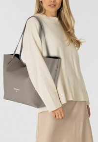 Bolso tote de cuero gris con superficie texturizada y abertura superior, con un logo minimalista. Llevado sobre un suéter crema y una falda beige.