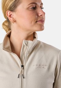Veste en polaire beige clair avec col montant, fermeture éclair et logo de montagne discret. Comprend un tissu texturé et un cordon de serrage ajustable.