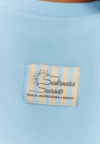 Světle modrá látka trička s tkanou etikou s béžovými a bílými pruhy a textem "Saltwater Sunsets" v černém písmu.