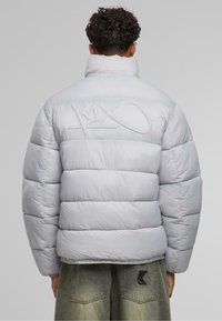 Jachetă puffer gri deschis, cu guler înalt, panouri cusute și detaliu brodat cu logo pe spate. Textură de material neted și lucios.