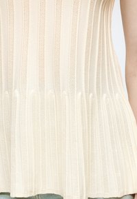 Gros plan d'un haut en maille côtelée couleur crème avec des rayures texturées verticales, porté avec un jean bleu clair.