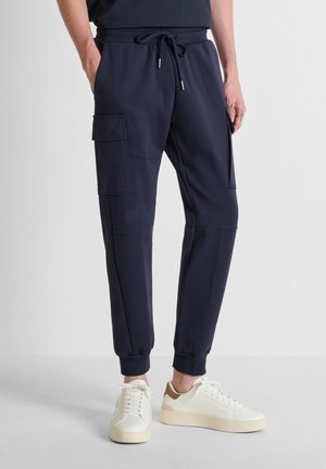 Uomo che indossa pantaloni cargo jogger blu navy e scarpe da ginnastica bianche, in piedi con le mani in tasca su uno sfondo semplice.