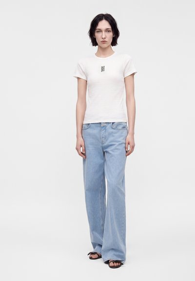 Jeune femme debout, portant un t-shirt blanc à manches courtes avec un petit logo et un jean large bleu clair avec des sandales noires.