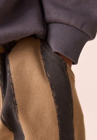 Braun-schwarz gestreifter Stoff mit strukturierter Oberfläche. Graue Bündchen eines Sweatshirts sind oberhalb des Bundes sichtbar. Detail zeigt die Naht.