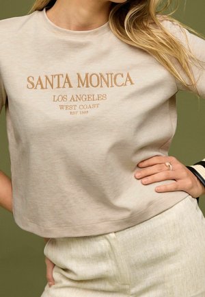 Beige cropped T-shirt met de tekst "Santa Monica Los Angeles West Coast Est 1988", gedragen met lichtgekleurde broek en hand op de heup.