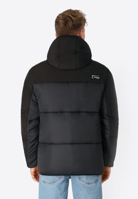 Schwarze Pufferjacke mit hohem Kragen und gesteppter Optik. Verfügt über einen einfarbig schwarzen Oberbereich und einen Logo-Patch auf der linken Schulter.
