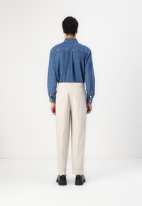 Camicia di jeans blu scuro con dettagli in bottoni sui polsini, abbinata a pantaloni beige su misura e scarpe nere. Sfondo neutro.