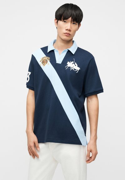 Νεαρός άνδρας που φοράει μπλε ναυτικό πόλο πουκάμισο με ανοιχτό μπλε διαγώνια ρίγα, λευκό λογότυπο παίκτη πόλο και λευκό παντελόνι, στέκεται μπροστά σε απλό φόντο.