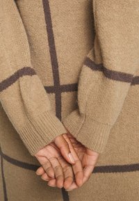 Un primo piano delle mani di una persona appoggiate dietro la schiena, che indossa un maglione beige a trama con motivi a scacchi marroni.