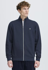 Marineblå zip-sweater laget av bomullsmiks med høy krage, lange ermer og en liten hvit logo på brystet. Myk tekstur.