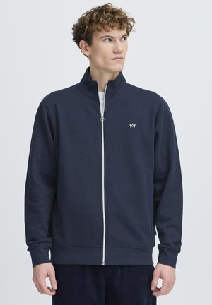 Marineblå zip-sweater laget av bomullsmiks med høy krage, lange ermer og en liten hvit logo på brystet. Myk tekstur.