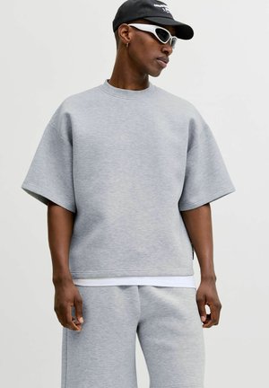 Mężczyzna w jasno szarej, oversize krótkiej bluzie z długim rękawem i pasujących spodniach, czarnej czapce i białych sportowych okularach przeciwsłonecznych, patrzy na bok.