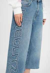 Culotte din denim de culoare albastră deschisă, având detalii uzate și un text brodat alb îndrăzneț pe lateral. Design cu pantaloni largi și tiv brut.