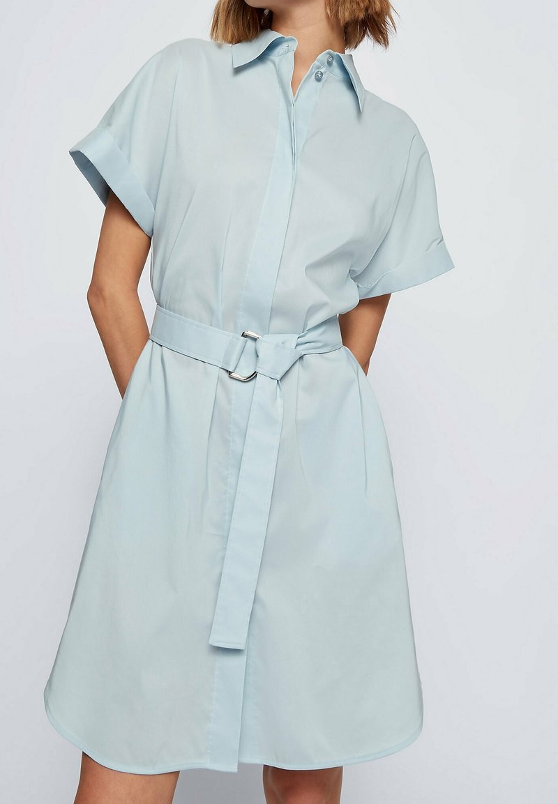 Jasnoniebieska sukienka koszulowa z krótkimi rękawami, z przyciskiem z przodu i paskiem w talii. Miękka tkanina i zaokrąglony dół. Płaski kołnierz i minimalistyczny design.