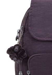 Lila tygryggsäck med klafföppning, dragkedjefickor och en fluffig gorillanyckelring fäst vid dragkedjan.